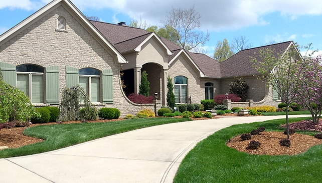 Large suburban house made of gray bricks that recently received landscape design service in the front yard featuring a sidewalk and small tree.