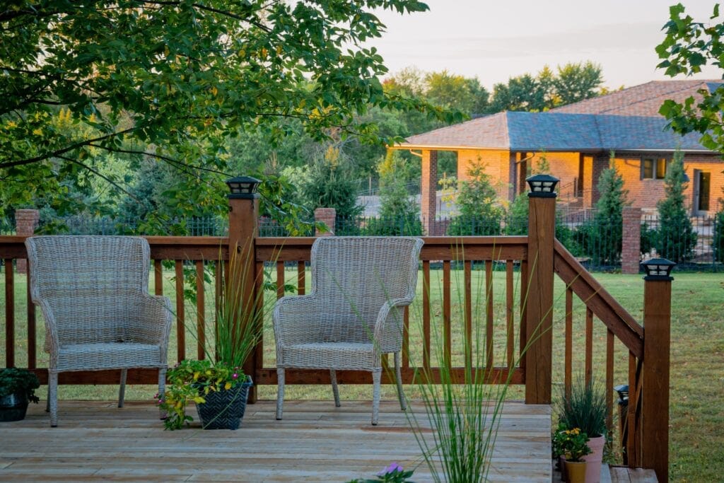 Two white wicker chairs sit on a wooden deck surrounded by potted plants, overlooking fresh spring landscaping in a grassy yard with trees and a brick house. The calm scene is bathed in soft evening light.