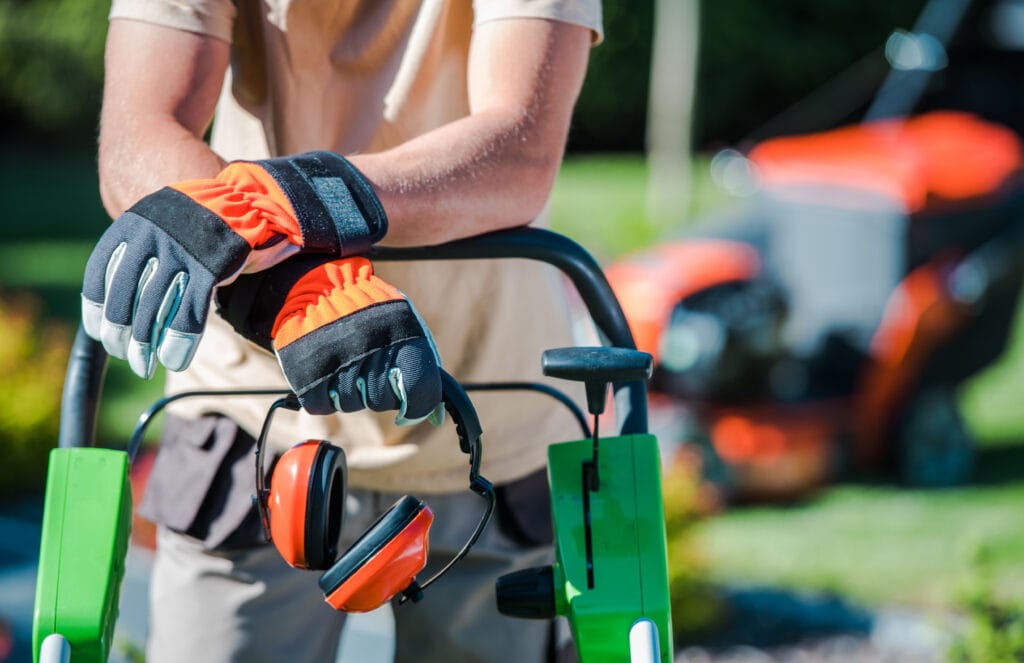Landscape professional wearing orange gloves and holding orange ear covers with hands resting on piece of landscaping equipment.