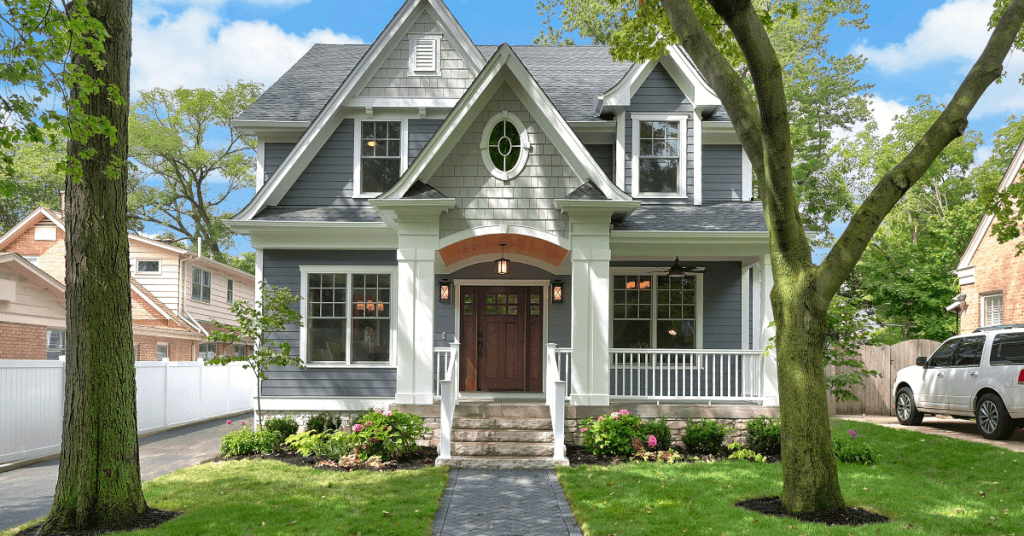 A small, gray Craftsman style home with white trim framed by two mature trees with a small but tidy flower bed out front, an example of a home with a landscape requiring affordable lawn service costs.