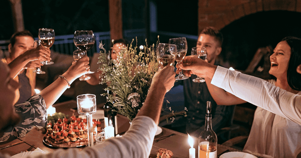 A group of people sit around a candlelit dinner table, raising their glasses in a toast and smiling. The table is decorated with flowers, candles, and plates of food. The atmosphere is warm and festive, and they are enjoying the new fall landscaping ideas they brought to life.