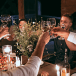 A group of people sit around a candlelit dinner table, raising their glasses in a toast and smiling. The table is decorated with flowers, candles, and plates of food. The atmosphere is warm and festive, and they are enjoying the new fall landscaping ideas they brought to life.