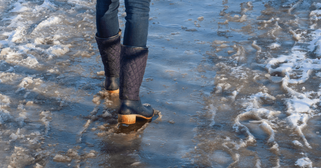 A person wearing dark jeans and black boots walks on a partially melted, icy and slushy path, with patches of snow and ice visible on the ground, a reminder of the importance of snow and frost protection.