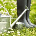 A pair of black rubber boots stands on grass among daisies, next to a metal watering can and several small metal containers, evoking a spring yard cleanup scene.