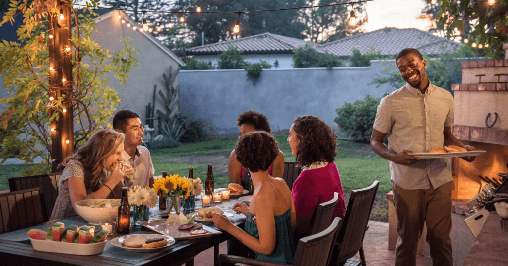 Six people enjoy an outdoor dinner at a backyard table decorated with flowers and string lights. One man stands and serves food while the others sit, talk, and laugh as evening sets in, showing beautiful landscape designs for small backyards.
