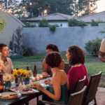 Six people enjoy an outdoor dinner at a backyard table decorated with flowers and string lights. One man stands and serves food while the others sit, talk, and laugh as evening sets in, showing beautiful landscape designs for small backyards.