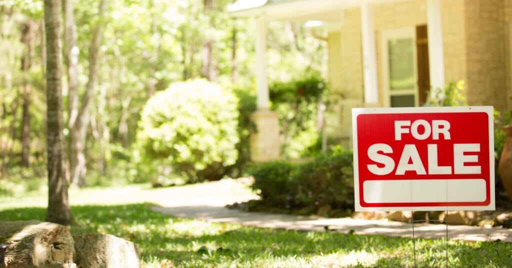 A red and white For Sale sign is posted in the front yard of a house with a green lawn and trees, with the house partially visible in the background. Homeowners are wondering, "does hardscape increase home value?" and "does landscaping increase home value?"