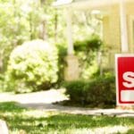 A red and white For Sale sign is posted in the front yard of a house with a green lawn and trees, with the house partially visible in the background. Homeowners are wondering, "does hardscape increase home value?" and "does landscaping increase home value?"