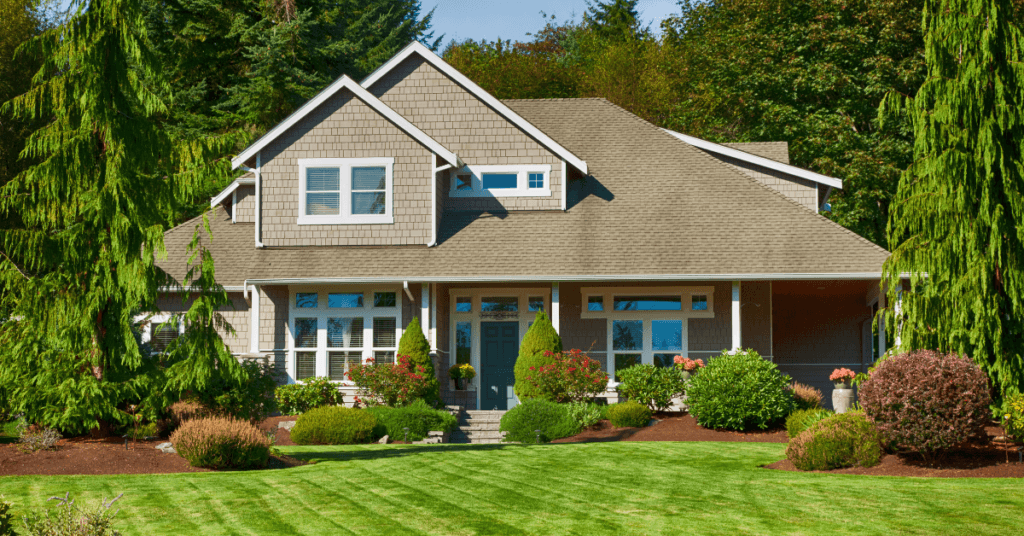A two-story suburban house with gray siding, white trim, and a neatly manicured lawn showcases beautiful neighborhood hoa landscaping, surrounded by lush green trees and landscaped bushes on a sunny day.